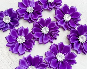 May include: A collection of ten vibrant purple satin flower embellishments. Each flower features layered petals and a sparkling rhinestone center. The flowers are arranged on a light-colored wooden surface, creating a visually appealing display.