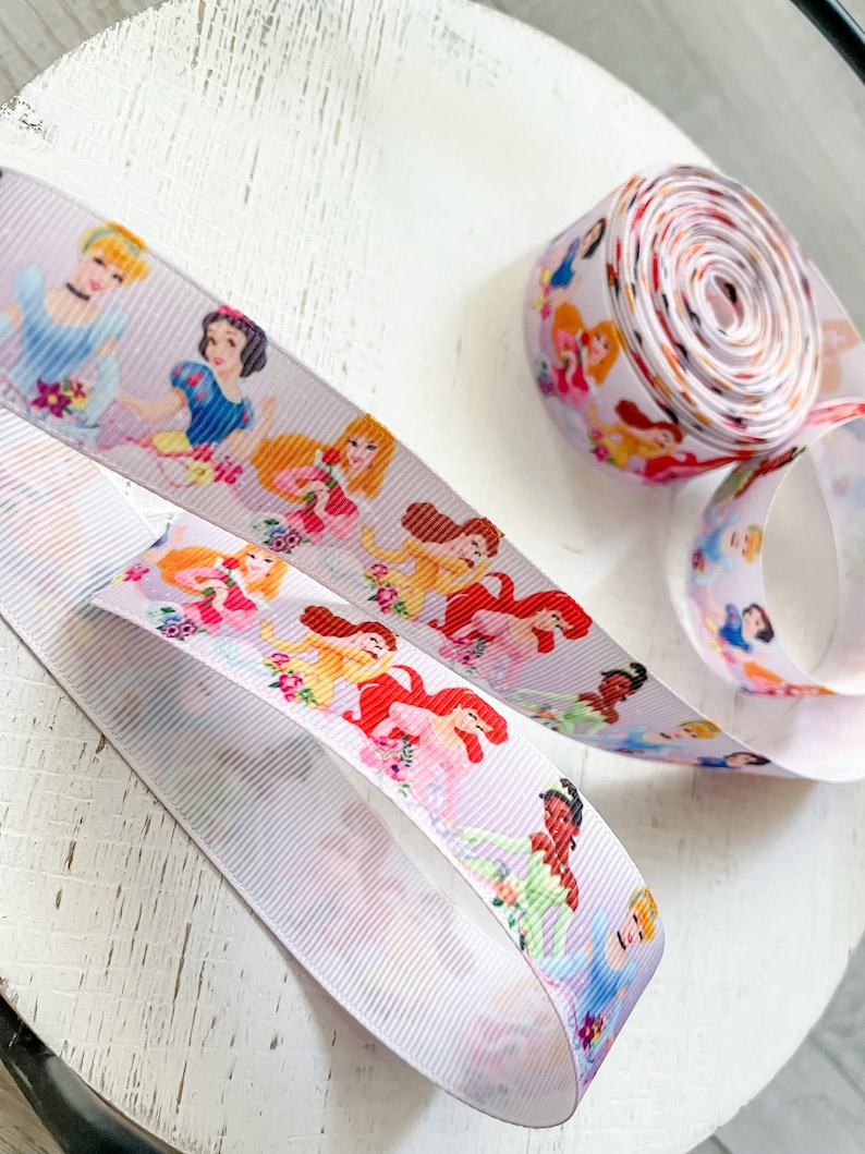 Puede incluir: Cinta blanca con ilustraciones coloridas de princesas de Disney. La cinta se muestra enrollada y desenrollada, mostrando a los personajes en varias poses. Ideal para manualidades, envolver regalos o a&ntilde;adir un toque m&aacute;gico a cualquier proyecto.