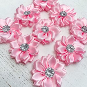 Peut inclure: Dix fleurs en ruban de satin rose clair, chacune avec un centre en strass argenté, sont disposées sur une surface en bois blanc. Ces fleurs décoratives sont idéales pour embellir les créations artisanales et les accessoires.