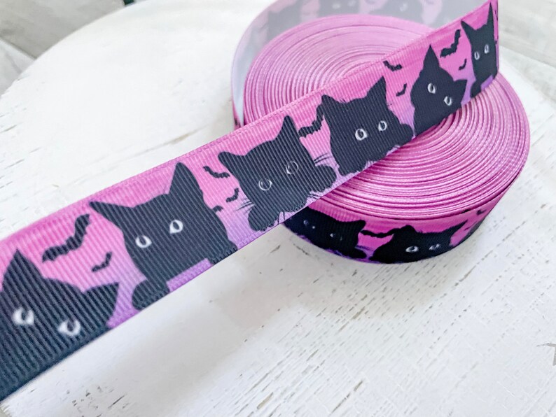 K&ouml;nnte beinhalten: Eine Rolle lila Band mit einem sich wiederholenden Muster aus schwarzen Katzen-Silhouetten und Flederm&auml;usen. Das Band hat einen Farbverlauf, der von Schwarz zu Lila &uuml;bergeht. Dieses dekorative Band ist ideal f&uuml;r Halloween-Bastelarbeiten und Geschenkverpackungen.