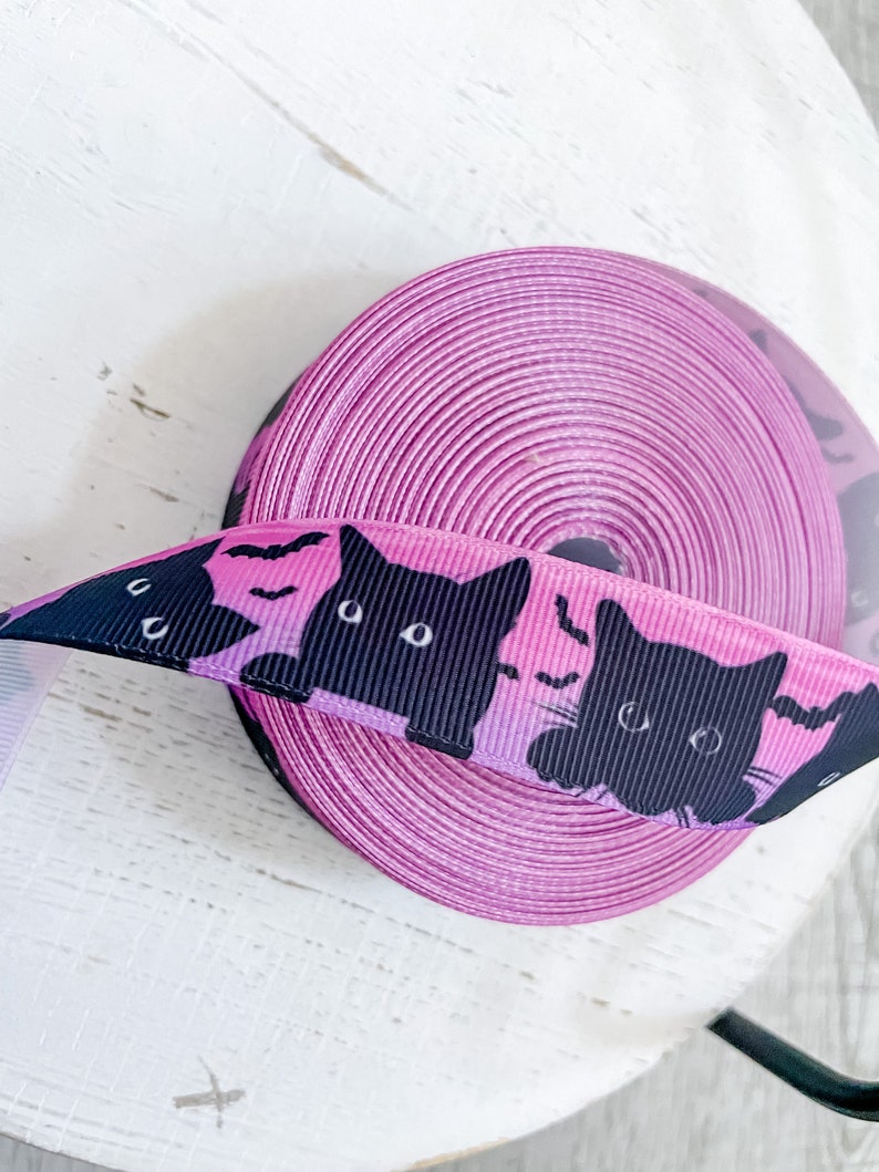 K&ouml;nnte beinhalten: Eine Rolle rosa Band mit einem Muster aus schwarzen Katzen und Flederm&auml;usen. Das abgerollte Band zeigt schwarze Katzensilhouetten und Flederm&auml;use vor einem rosa-violetten Farbverlauf. Geeignet f&uuml;r Bastelarbeiten oder zum Geschenkeverpacken.