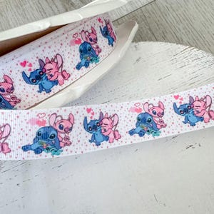 May include: White ribbon featuring a repeating pattern of blue and pink cartoon characters with hearts. The ribbon is in a white box and is displayed on a light wood surface. The ribbon is suitable for crafting and gift wrapping.