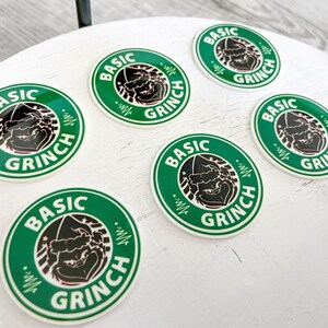 May include: Circular green and white stickers with the text "BASIC GRINCH" in a circular design. The stickers feature a black illustration of the Grinch character. The stickers are arranged on a white surface.