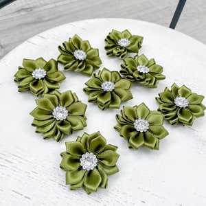 May include: Olive green satin flower embellishments with a sparkling rhinestone center. These decorative flowers are arranged on a white surface. The flowers have a layered petal design.