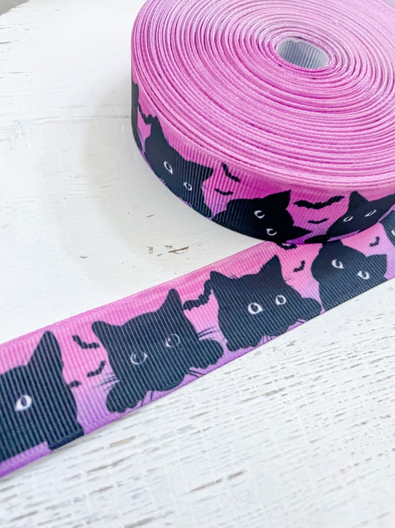K&ouml;nnte beinhalten: Eine Rolle rosa Band mit einem sich wiederholenden Muster aus schwarzen Katzen-Gesichtern und Flederm&auml;usen. Das Band hat einen leuchtenden Rosaton, der zu einem helleren Farbton verblasst und einen Farbverlauf erzeugt. Das Design mit schwarzen Katzen und Flederm&auml;usen ist ideal f&uuml;r Halloween-Bastelarbeiten.