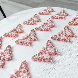 Op de afbeelding: Roze vlinderversieringen met zilveren strass-steentjes. Deze decoratieve vlinderaccenten zijn gerangschikt op een wit oppervlak. De vlinders hebben een verhoogd, gestructureerd ontwerp en zijn ideaal voor knutselprojecten.