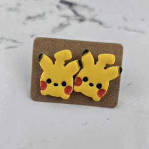 May include: A pair of yellow Pikachu stud earrings with black accents and red cheek markings. The earrings are displayed on a brown cardboard backing.