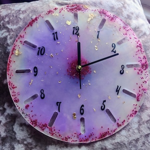 May include: A purple and white resin clock with gold glitter and pink glitter. The clock face has black numbers 1 through 12 and black hour and minute hands.