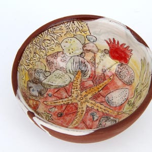 May include: A round ceramic bowl with a terracotta exterior and a white interior featuring a hand-painted marine scene. The design includes a yellow starfish, various grey and brown pebbles, coral-like structures, and a bright red sea anemone.