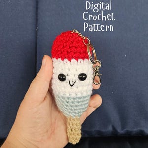 May include: A crochet popsicle keychain featuring a red top, white middle, and light blue bottom section with a tan stick. The popsicle has two black bead eyes and a stitched black V-shaped mouth. The text Digital Crochet Pattern is visible.