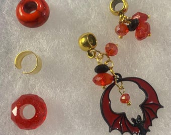 May include: A collection of jewelry components, including red beads, a gold-colored ear cuff, and gold-toned charms with red and black crystal accents. One charm features a red bat design.