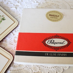 May include: A white box with a red and black stripe. The box has the text "Pimpernel De Luxe Finish" and a sticker that says "Marzolf Idstein".