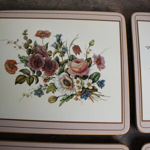 K&ouml;nnte beinhalten: Set aus vier quadratischen Tischsets mit Blumendesigns. Jedes Tischset hat einen cremefarbenen Hintergrund mit einem Blumenstrau&szlig; in Rosa-, Rot-, Wei&szlig;- und Blaut&ouml;nen. Die R&auml;nder sind mit einem rosa Rand und einem goldenen Akzent versehen.