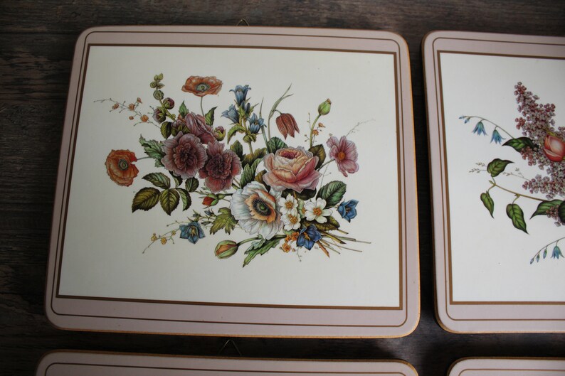 K&ouml;nnte beinhalten: Set aus vier quadratischen Tischsets mit Blumendesigns. Jedes Tischset hat einen cremefarbenen Hintergrund mit einem Blumenstrau&szlig; in Rosa-, Rot-, Wei&szlig;- und Blaut&ouml;nen. Die R&auml;nder sind mit einem rosa Rand und einem goldenen Akzent versehen.