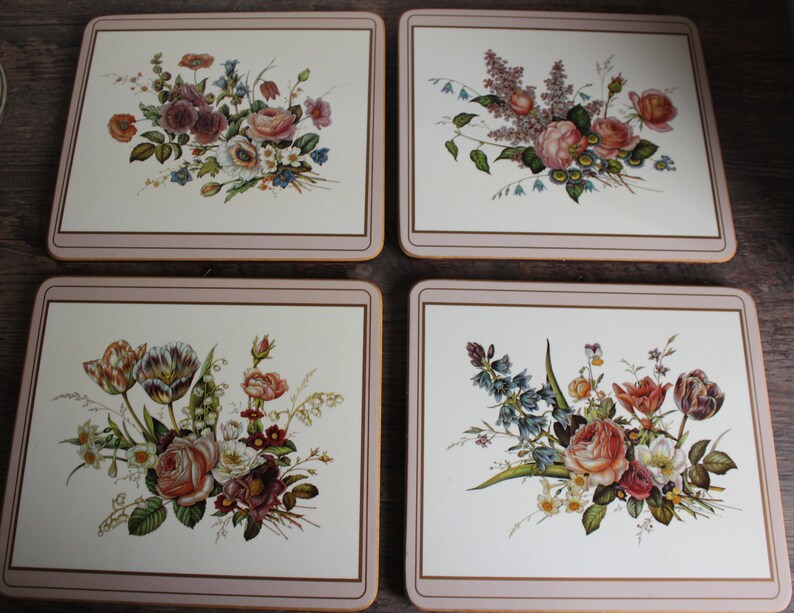 K&ouml;nnte beinhalten: Set aus vier quadratischen Tischsets mit Blumendesigns. Jedes Tischset hat einen cremefarbenen Hintergrund mit einer anderen Anordnung von Rosen, Tulpen und anderen Blumen in Rosa-, Rot- und Lilat&ouml;nen. Die R&auml;nder sind mit einem hellbraunen Rand versehen.