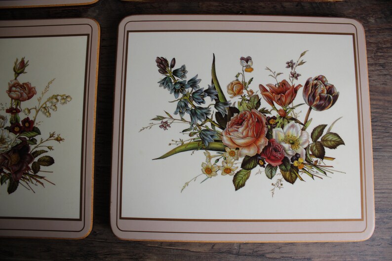 K&ouml;nnte beinhalten: Rechteckige Tischsets mit floralen Designs. Die Tischsets haben einen cremefarbenen Hintergrund mit einem Rand und zeigen einen Strau&szlig; aus Rosen, Tulpen und anderen Blumen in Rot-, Orange- und Wei&szlig;t&ouml;nen.