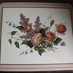 K&ouml;nnte beinhalten: Quadratischer Platzdeckchen mit floralem Design. Der Platzdeckchen zeigt einen Strau&szlig; aus rosa und pfirsichfarbenen Rosen, blauen Vergissmeinnicht und Fliederzweigen auf wei&szlig;em Hintergrund. Der Rand ist hellrosa mit einer d&uuml;nnen goldenen Linie.