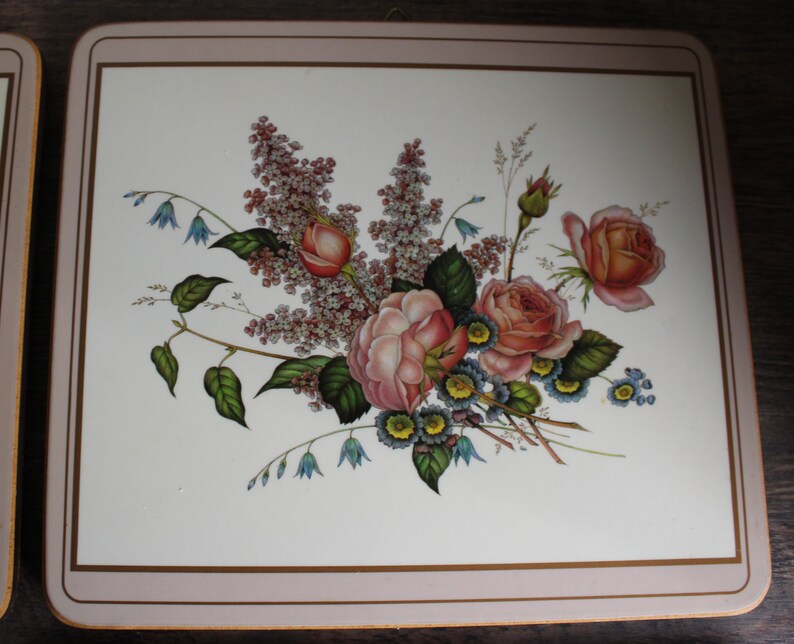 K&ouml;nnte beinhalten: Quadratischer Platzdeckchen mit floralem Design. Der Platzdeckchen zeigt einen Strau&szlig; aus rosa und pfirsichfarbenen Rosen, blauen Vergissmeinnicht und Fliederzweigen auf wei&szlig;em Hintergrund. Der Rand ist hellrosa mit einer d&uuml;nnen goldenen Linie.