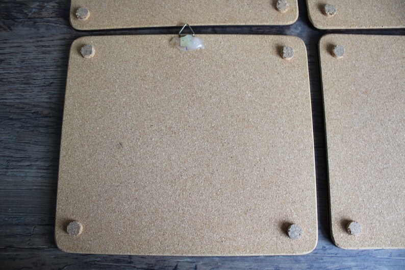 K&ouml;nnte beinhalten: Ein Satz von vier quadratischen Korktafeln mit abgerundeten Ecken. Jede Tafel hat eine Korkr&uuml;ckseite und kleine, runde Korkf&uuml;&szlig;e. Eine Tafel hat einen Metallb&uuml;gel auf der R&uuml;ckseite. Die Tafeln sind auf einer dunklen Holzoberfl&auml;che angeordnet.