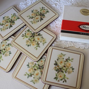 May include: Set of six square coasters with a floral design featuring white roses. The coasters are white with a gold trim and are packaged in a white box with the text "Pimpernel De Luxe Finish" and "Marzolf Gosten" printed on the box.