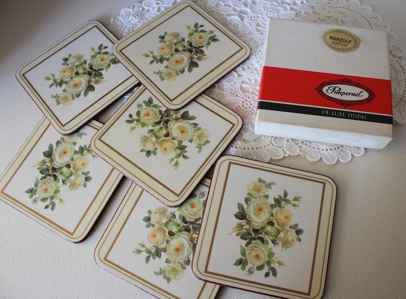 May include: Set of six square coasters with a floral design featuring white roses. The coasters are white with a gold trim and are packaged in a white box with the text "Pimpernel De Luxe Finish" and "Marzolf Gosten" printed on the box.