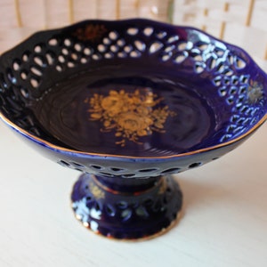 Op de afbeelding: Een blauwe en gouden keramische fruitschaal met een decoratief ontwerp. De schaal heeft een voet en een tralieontwerp rond de rand.