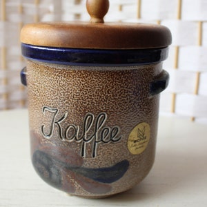 Könnte beinhalten: Eine braune Kaffeedose aus Keramik mit einem Holzdeckel. Die Dose hat ein blaues und braunes Blumenmuster und das Wort "Kaffee" auf der Vorderseite. Die Dose hat einen goldenen Aufkleber mit dem Wort "Scholz" darauf.