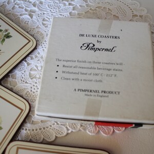 May include: A box of white ceramic coasters with a floral design. The box is labelled "De Luxe Coasters by Pimpernel". The coasters are made in England and are heat resistant up to 100 degrees Celsius.
