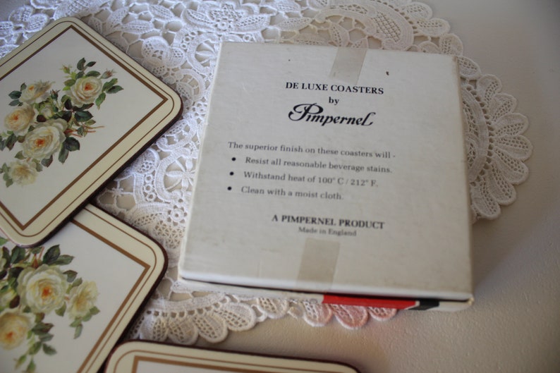 May include: A box of white ceramic coasters with a floral design. The box is labelled "De Luxe Coasters by Pimpernel". The coasters are made in England and are heat resistant up to 100 degrees Celsius.