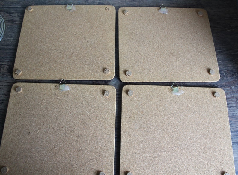 K&ouml;nnte beinhalten: Vier quadratische Korkplatten mit abgerundeten Ecken. Jede Platte hat einen kleinen Metallb&uuml;gel oben und vier kleine, runde Korkf&uuml;&szlig;e an den Ecken. Die Platten sind hellbraun.