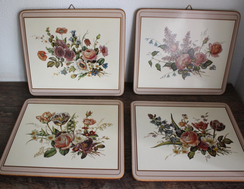 K&ouml;nnte beinhalten: Vier quadratische Tischsets mit Blumendesigns. Jedes Tischset hat einen cremefarbenen Hintergrund mit einer anderen Anordnung von Rosen, Tulpen und anderen Blumen in Rosa, Rot und Lila. Ein hellbrauner Rand umrahmt jedes Tischset.