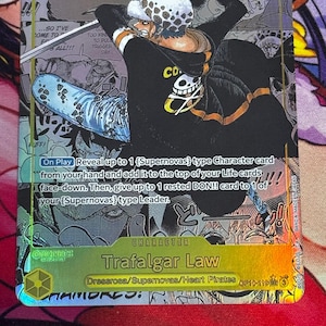 May include: A One Piece trading card featuring Trafalgar Law in a black hoodie with a skull emblem, wielding a sword. The card has a gold border, a power level of 9000, and text detailing the On Play ability to reveal Supernovas type character cards.