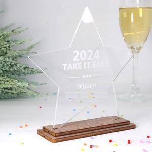 Può includere: Un trofeo a forma di stella in acrilico trasparente con la scritta "2024 TAKE IT EASY Winner". Montato su una base in legno a due livelli. Sullo sfondo, un bicchiere di champagne e coriandoli.
