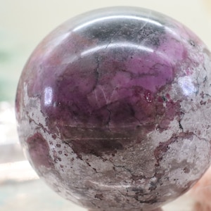 Puede incluir: Una esfera pulida de color púrpura y gris con un patrón de mármol. La esfera está hecha de piedra natural y tiene una superficie lisa y pulida.