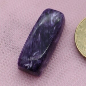Könnte beinhalten: Ein violetter und weißer Cabochon-Edelstein mit einem wirbelnden Muster. Der Edelstein ist etwa so groß wie eine deutsche 1-Euro-Münze.