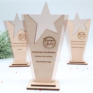 以下が含まれることがあります： 透明なアクリルサイドと白い星が付いた、木製のTeenager Awardトロフィーが3つ。トロフィーには、ドイツ語で「Teenager Award 2024」と受賞者の名前が刻まれています。ベースは正方形です。