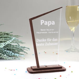 Può includere: Un trofeo in acrilico trasparente con cornice e base in legno scuro. Il trofeo presenta la parola "Papa" in bianco, insieme ad altro testo. Un bicchiere di champagne è sullo sfondo.
