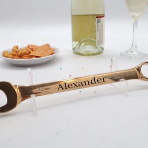May include: A bronze-coloured wrench with the name "Alexander" engraved on the handle. The wrench is 22mm in size. A bottle of wine, a wine glass, and a small plate of snacks are in the background.