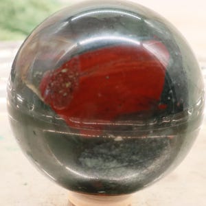 Könnte beinhalten: Eine polierte Edelstein-Kugel mit einem dunkelgrünen und roten Muster. Die Kugel ruht auf einem hellen Holzsockel. Der rote Bereich hat ein marmoriertes Aussehen.