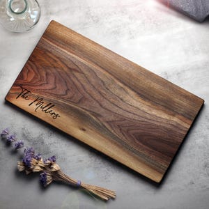 May include: A rectangular wooden cutting board with a natural wood grain pattern. The text "The Millers" is engraved on the board. A small bundle of dried lavender and a glass bottle are also visible.
