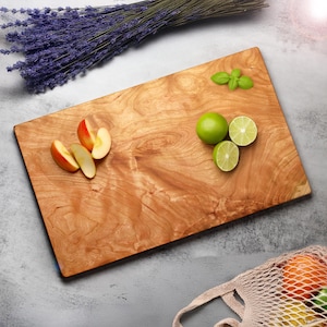May include: A rectangular wooden cutting board with a light brown finish. Sliced apples, limes, and basil are arranged on the board. A bundle of lavender and a mesh bag with oranges are also in the image.