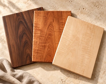 May include: Three rectangular wooden cutting boards arranged in a fan shape. The boards feature distinct wood grain patterns and colors, ranging from a dark brown, to a medium reddish-brown, and a light cream-colored wood finish.