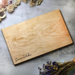 May include: A rectangular wooden cutting board with a light wood grain pattern. The words "Sara's Kitchen" are written in a cursive font on the lower left corner. The board is surrounded by dried flowers and greenery.