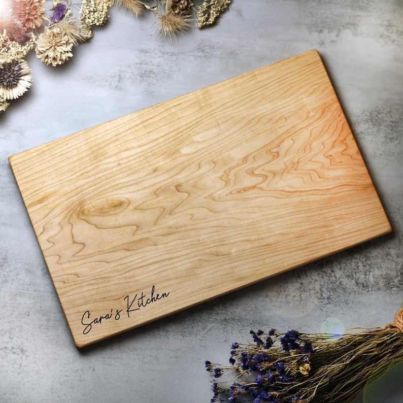 May include: A rectangular wooden cutting board with a light wood grain pattern. The words "Sara's Kitchen" are written in a cursive font on the lower left corner. The board is surrounded by dried flowers and greenery.