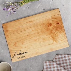 May include: A rectangular wooden cutting board with a light brown finish. The name "Anderson" and the text "Est. 2025" are printed in black script. A bundle of lavender and a checkered cloth are in the background.