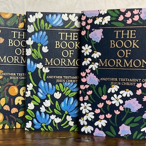 May include: Three hardback books titled "The Book of Mormon" with floral designs. The covers are navy blue with gold lettering and painted flowers in various colours, including blue, white, and pink. The text "Another Testament of Jesus Christ" is also visible.