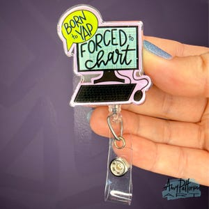 May include: A clear acrylic badge reel with a computer design. The design features a computer monitor with the text "BORN to YAP FORCED to chart" in black, green, and pink. The reel has a clear plastic strap and a metal clip.