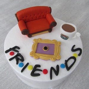 May include: A white cake decorated with a red couch, a purple picture frame, a coffee cup with the words "Central Perk" on it, and the word "FRIENDS" in black letters with colourful dots.