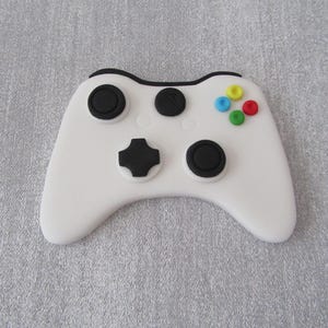 May include: A white games controller with black buttons and coloured accent buttons. The controller has a classic design, with a D-pad, analogue sticks, and action buttons. The controller is set against a grey background.