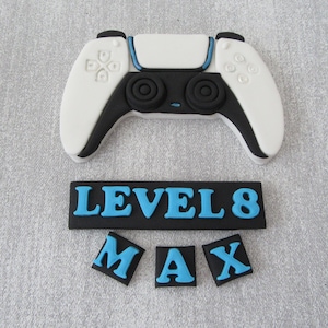 May include: A white and black video game controller with blue buttons. The controller is on a grey surface with a black banner that says "LEVEL 8" and three black squares with blue letters that spell "MAX".